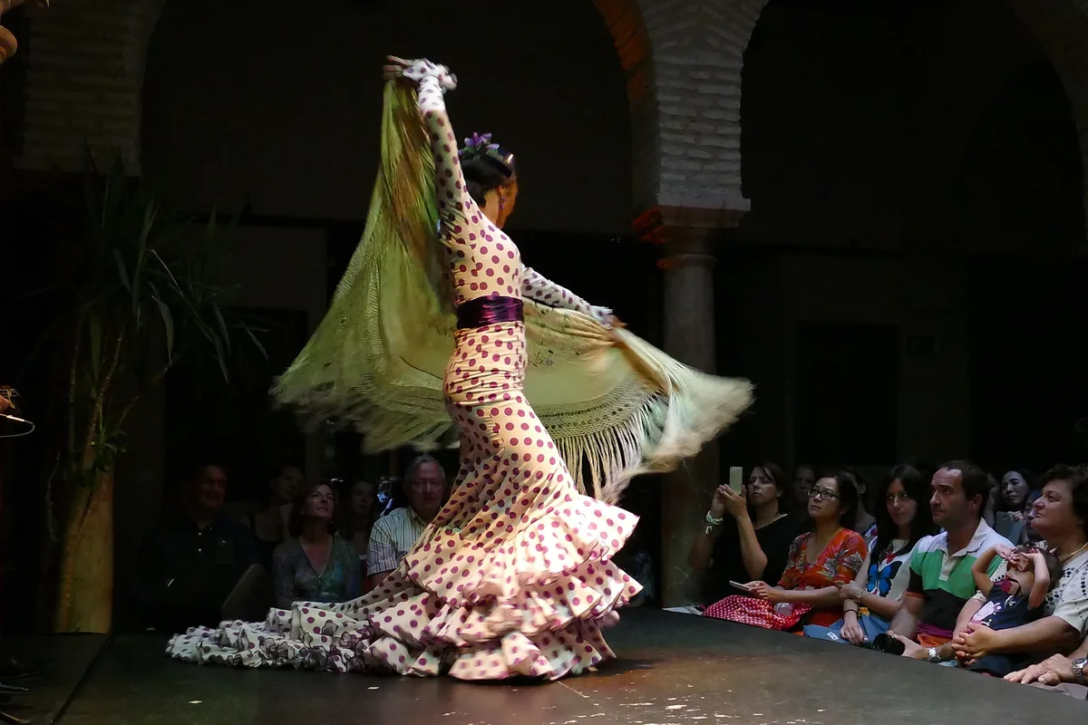 A flamenco dancer in a polka-dot dress turns with a long fringed shawl on a low stage, with seated spectators on the right and brick arches behind