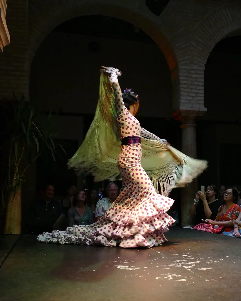 Una ballerina di flamenco in abito a pois ruota con una lunga scialle a frange sul palco basso del museo, con gli spettatori seduti sulla destra e archi in mattoni sullo sfondo