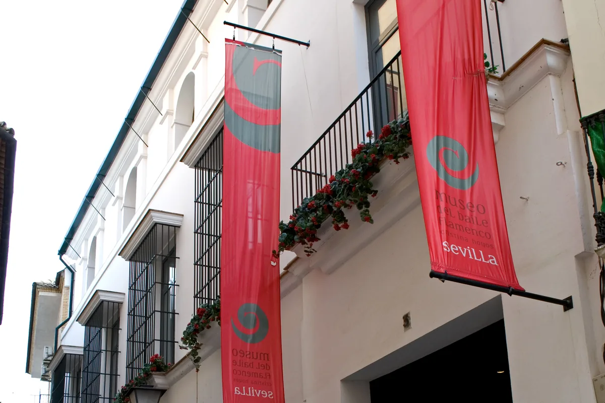La facciata bianca su strada del Museo del Baile Flamenco con striscioni rossi, un balcone di fiori rossi e una porta scura aperta