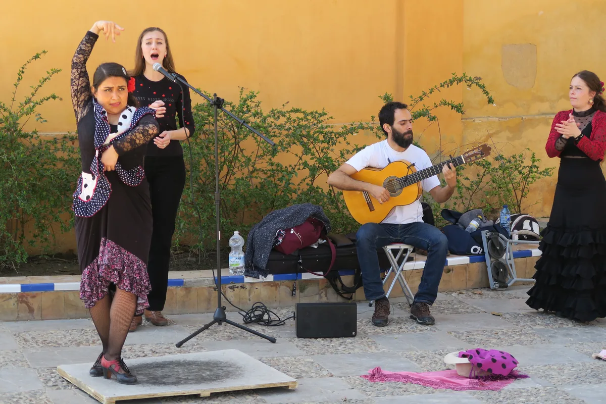 Una ballerina di flamenco, un cantante, un chitarrista e una seconda ballerina che batte le mani, si esibiscono su una piazza pavimentata di Siviglia davanti a un muro giallo